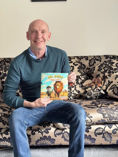Author Antony Flynn-Few sitting on a sofa holding a copy of Mr Jelby and the Lion